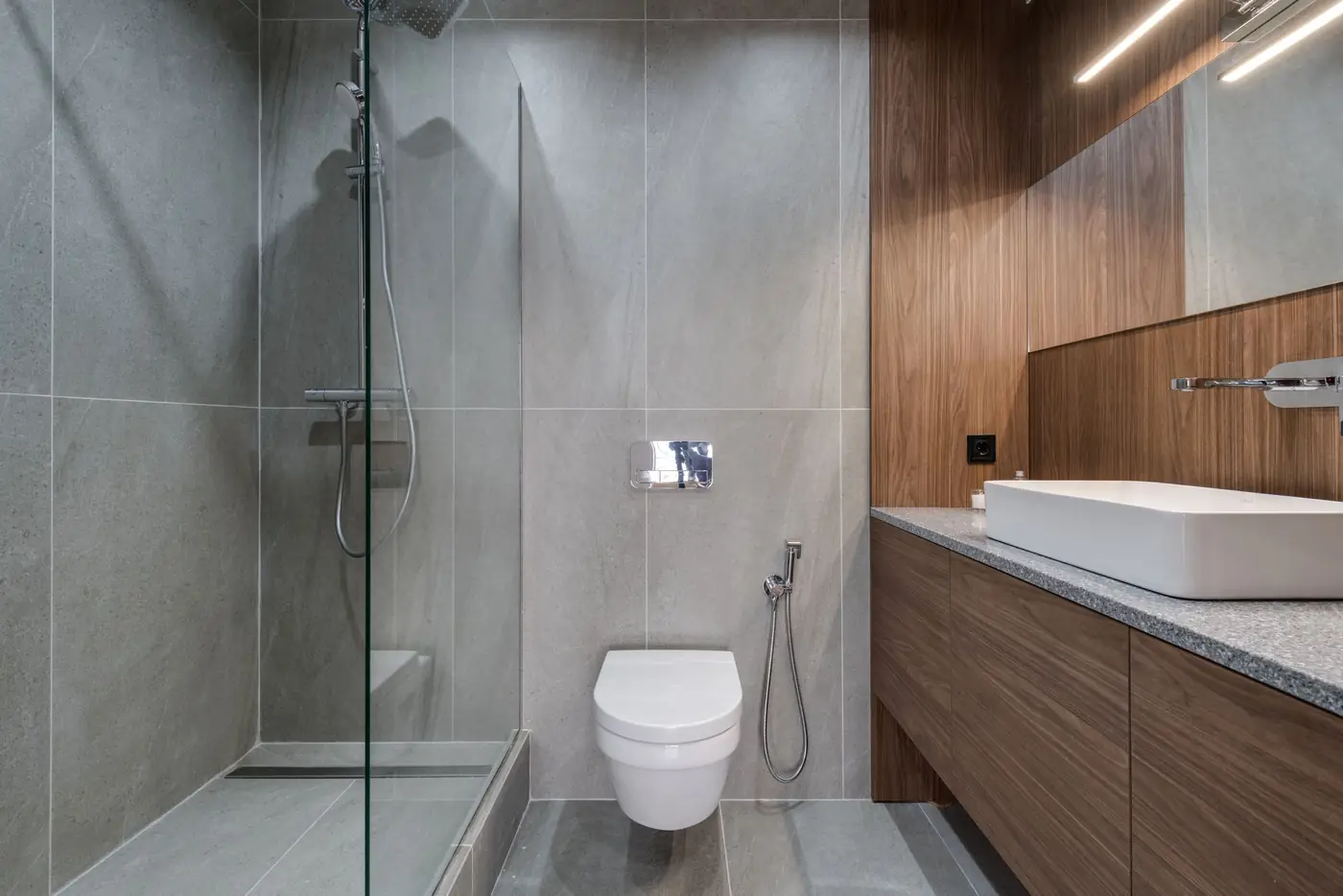 Badkamer met grijze tegels, inloopdouche in glas en een houten meubel