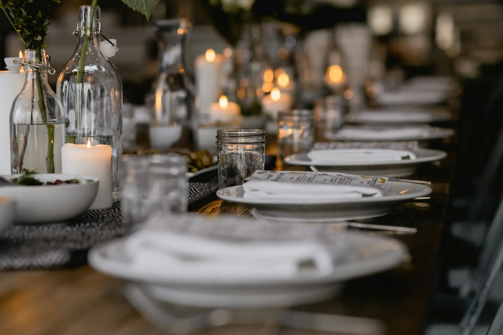 Lange tafel met brandende kaarsen, witte borden en glazen flessen