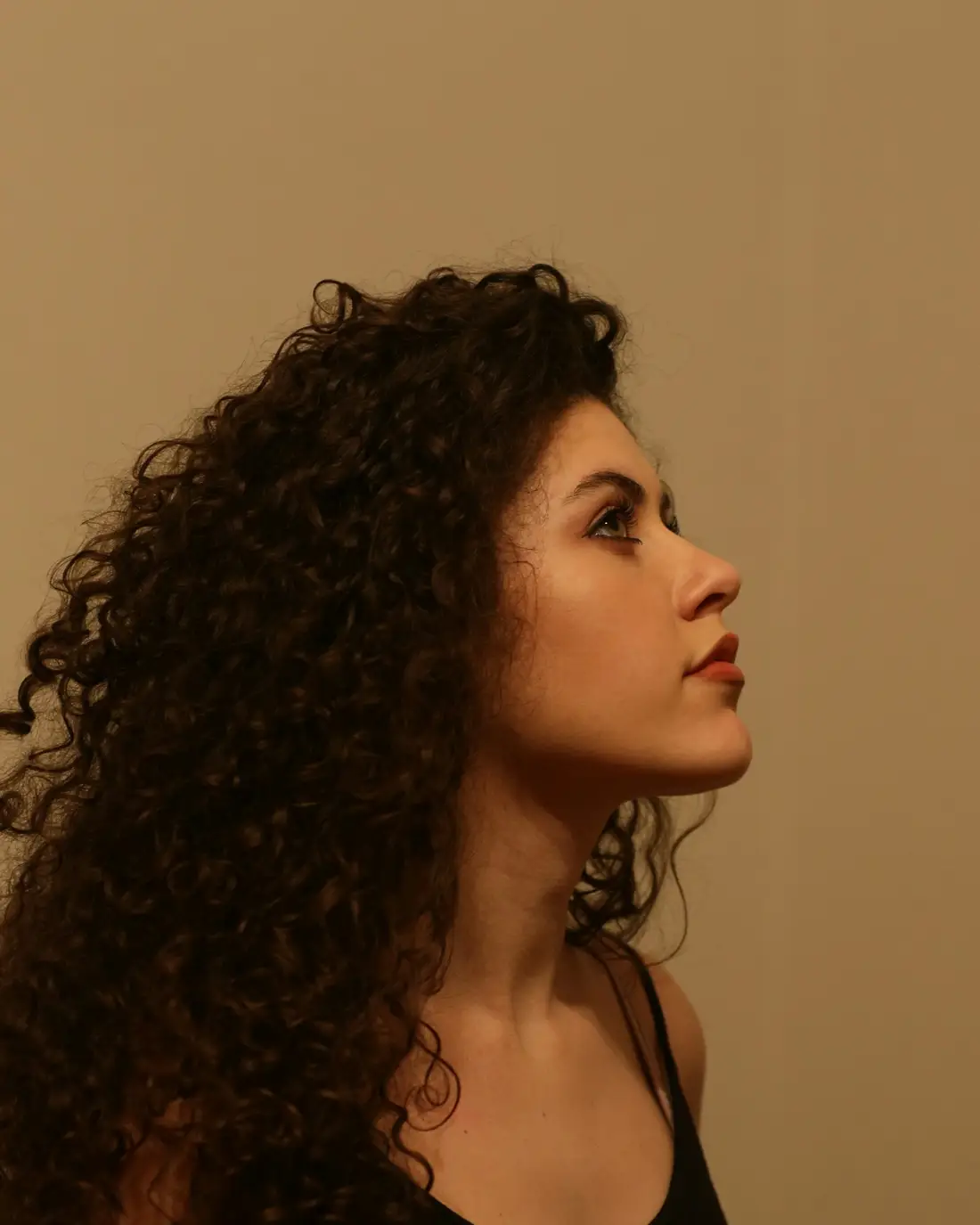 Een vrouw met volle donkere krullen kijkt opzij tegen een warme beige muur