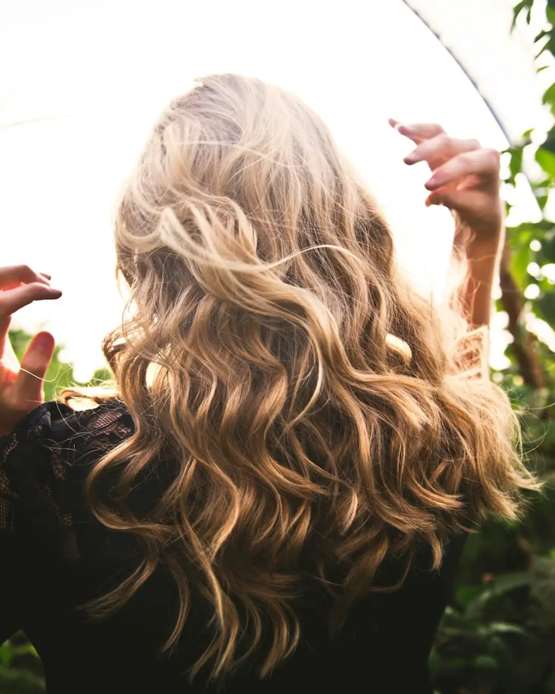 Een vrouw met lang golvend blond haar tussen groene planten, van achteren